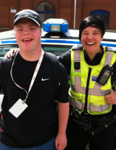 A person with learning disabilities smiling with a police officer in front of a police car